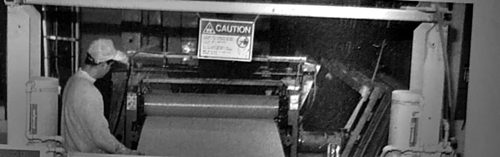 A man in a cap works at a laminating line, feeding a wide sheet through the rollers under a caution sign.
