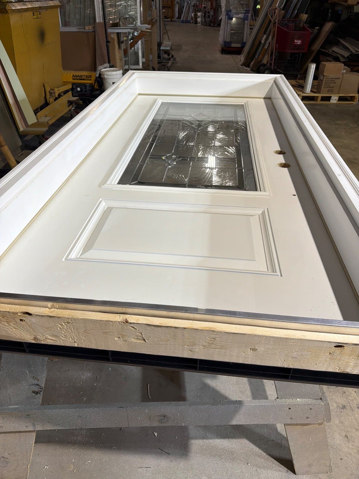 A white entry door slab lying flat on blocking in the shop, a rectangular clear lite above a single raised panel.