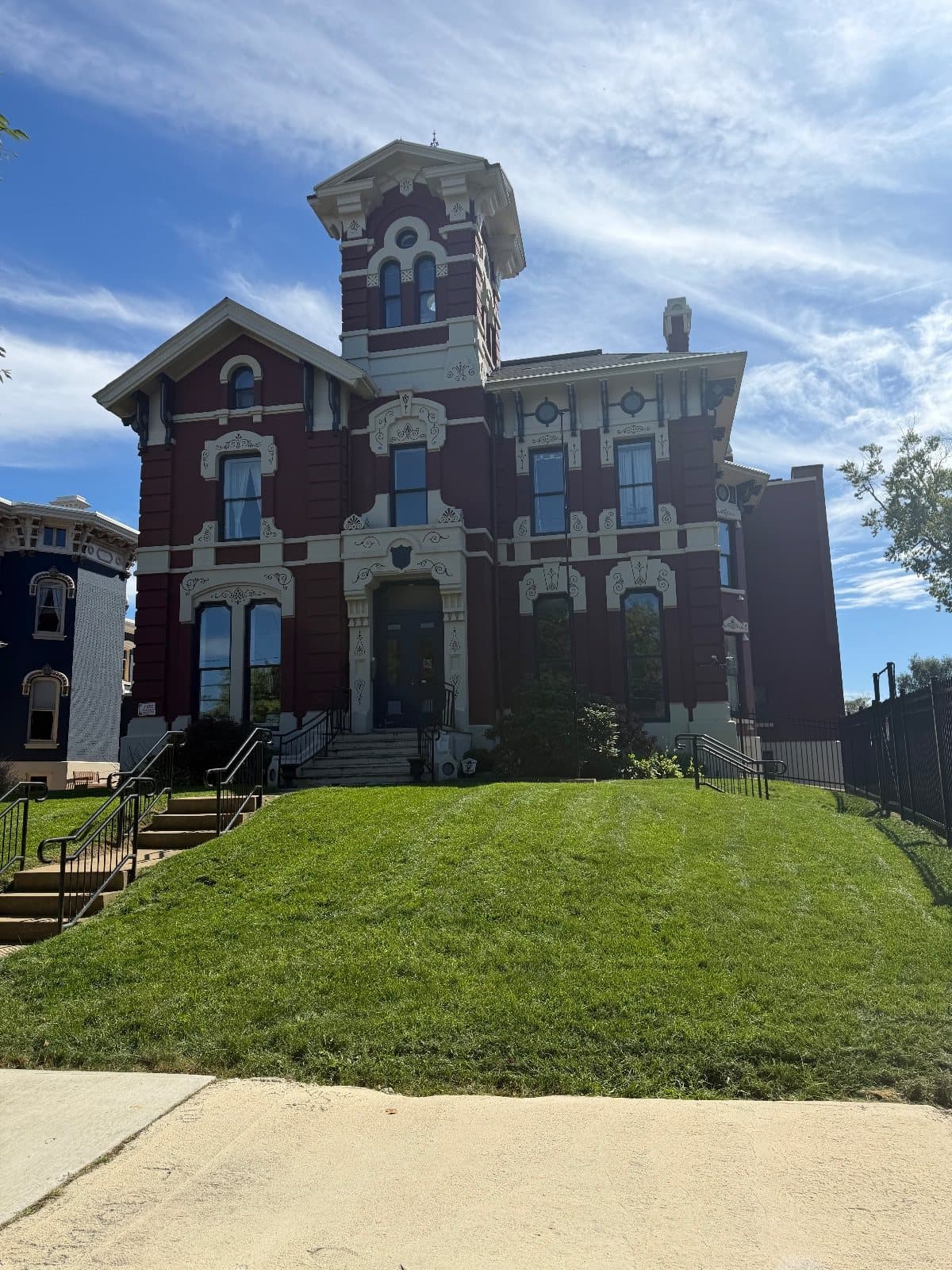 A red brick Italianate mansion with a square tower and cream stone window hoods, up a grass bank from the sidewalk.