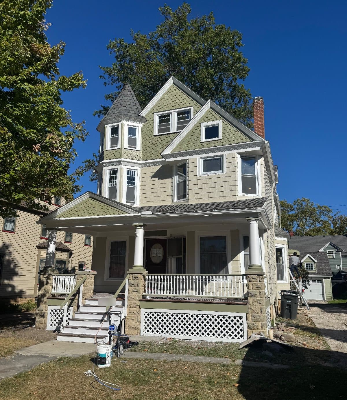 A three-story Victorian house in sage and cream shingle, a corner tower with a conical roof, two front gables and a stone-pillared porch, every window in a new white frame.