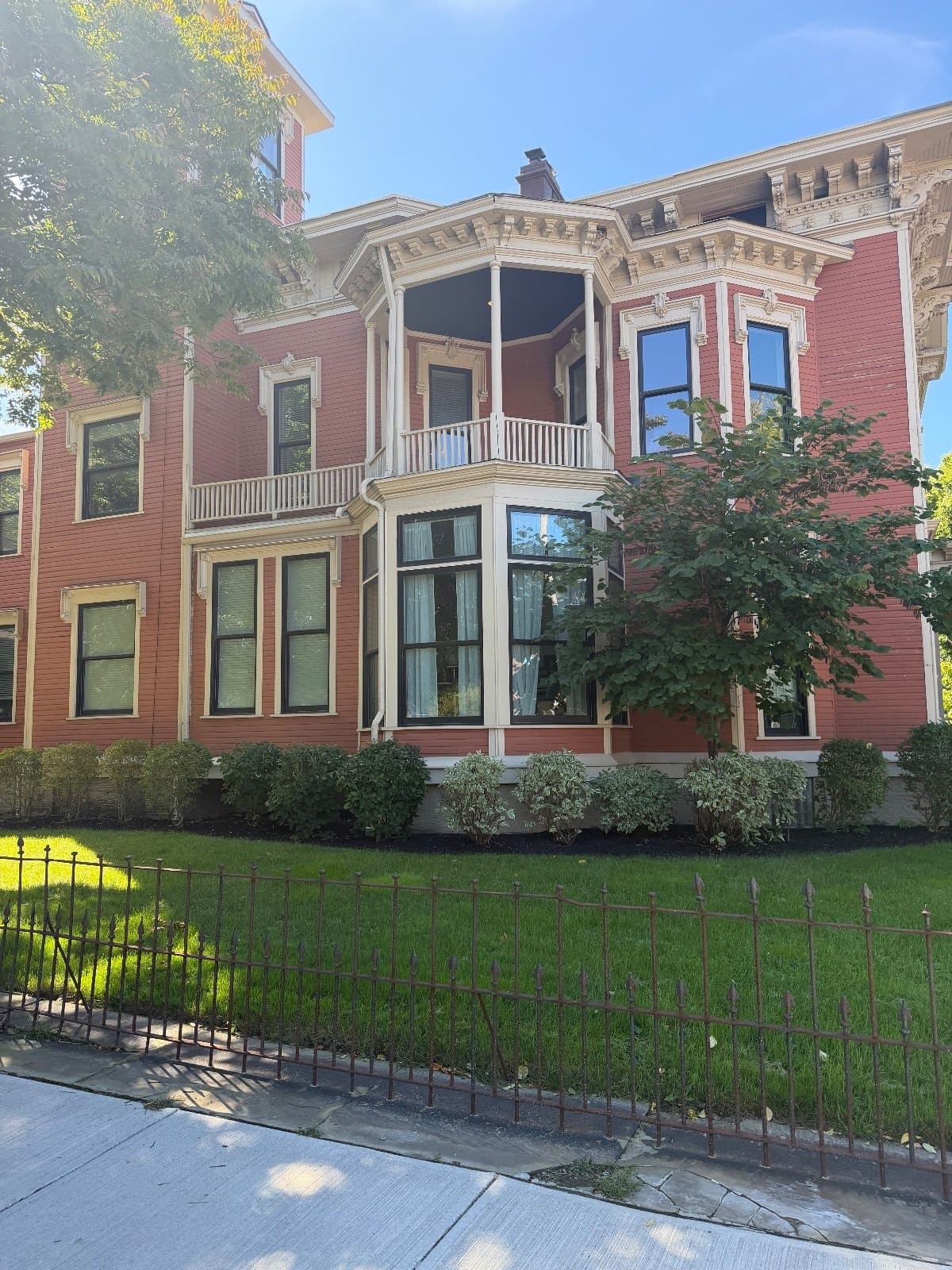 A red Victorian mansion with a two-story bay, a columned balcony above it and ornate white window hoods, behind an iron fence.