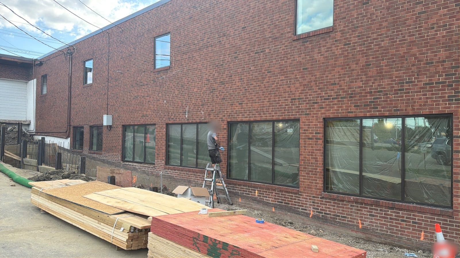 A long two-story red brick commercial building, a run of wide fixed picture windows in dark aluminum frames along the ground floor with protective film still on the glass, single windows on the upper floor, a man on a stepladder at the wall and stacked lumber in front.