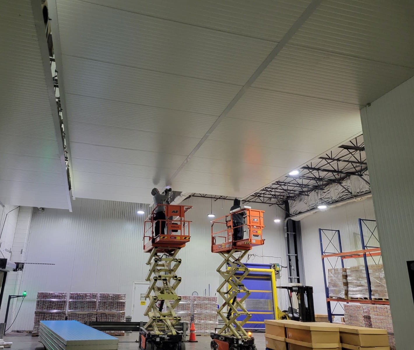 A room lined with white insulated metal panels, two workers on scissor lifts at the ceiling, a high-speed roll-up door and pallets of product behind them, a stack of new panels on the floor.