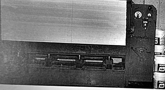 A panel press with a finished laminated panel held across its bed, gauges on the control column and adhesive pails stacked alongside.