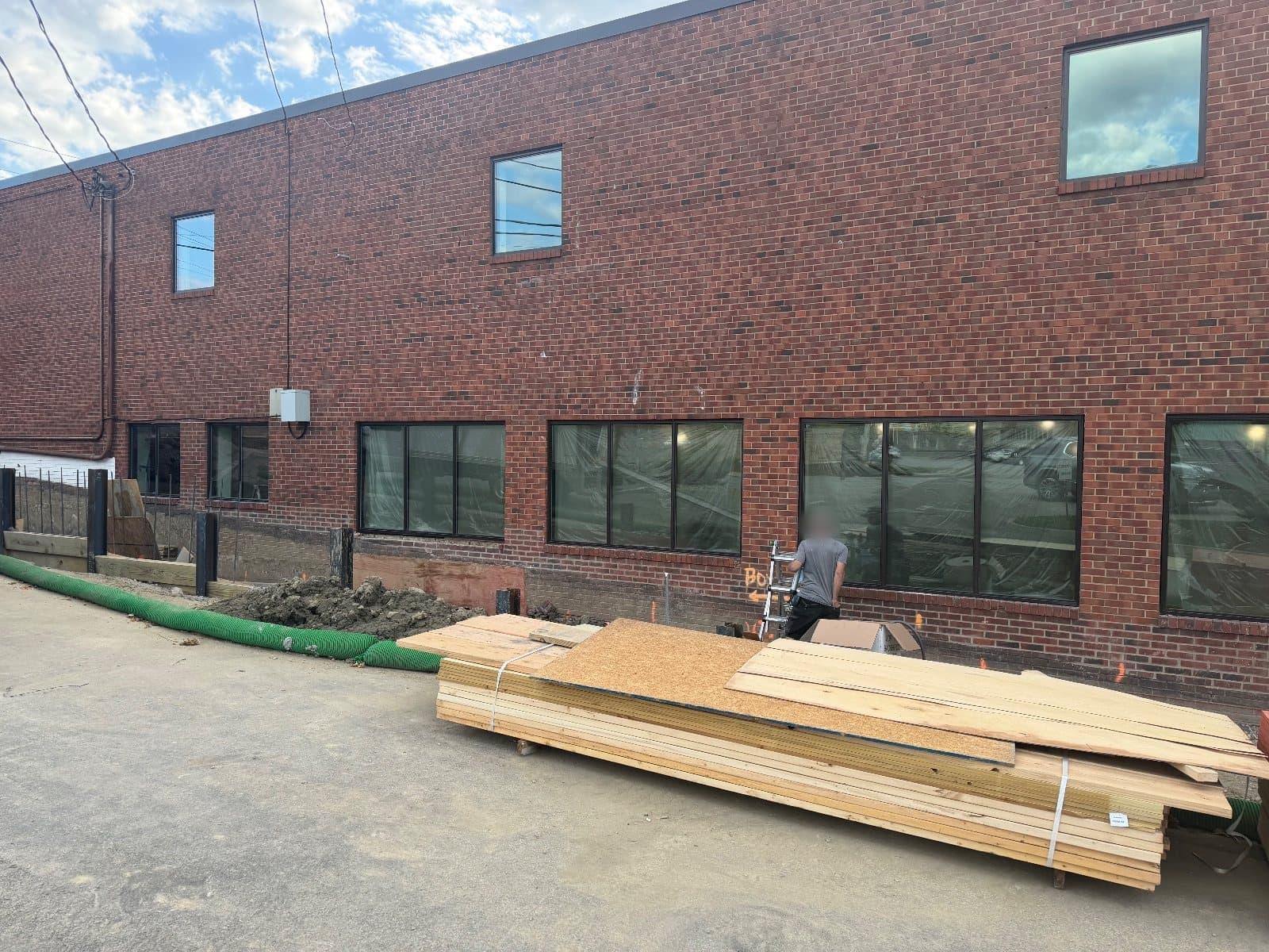 The same brick elevation from further along, four ground-floor runs of picture windows in dark aluminum frames, three single windows above, a stack of lumber on the pavement.