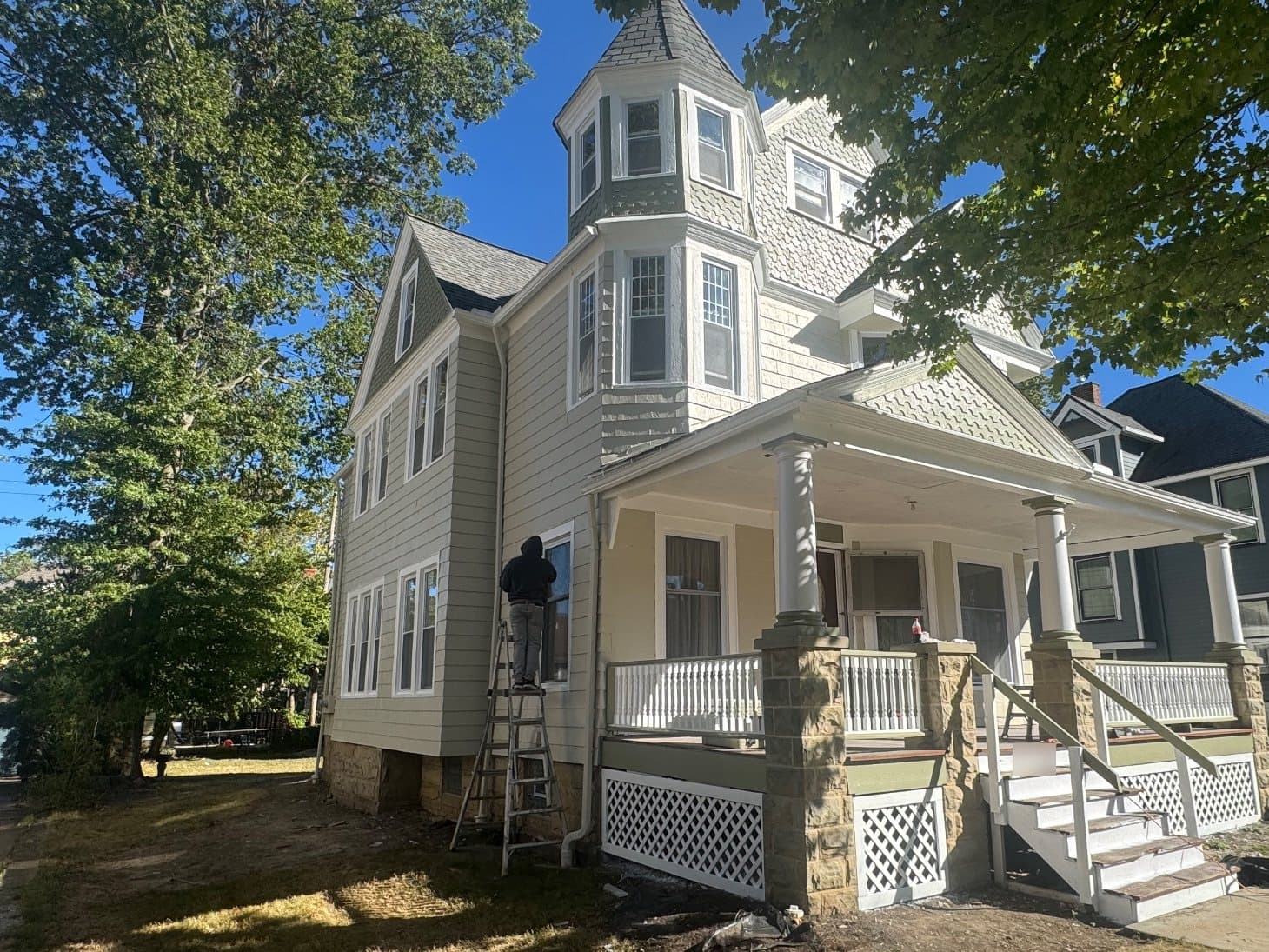 The same Victorian from the front corner, a man on a stepladder at a side window, the corner tower and a deep wraparound porch.