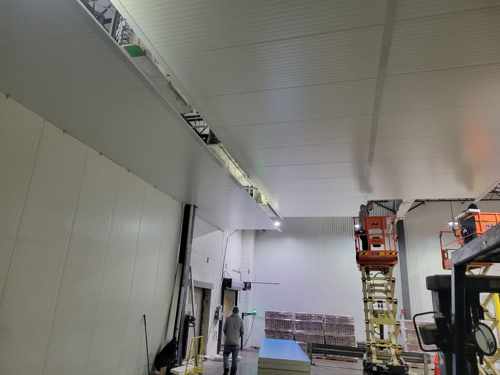 A long white insulated panel wall running the length of the room, a worker on a scissor lift at the far end, panels stacked on the floor.