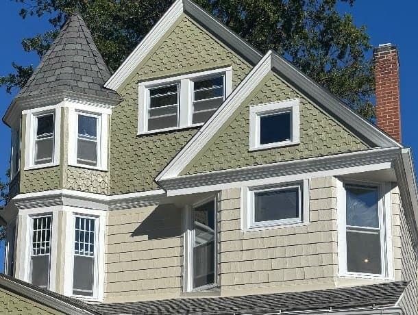 Close view of the upper floors: the corner tower's windows, a pair of double-hung windows in the front gable and a single window in the smaller gable, all in white frames against fish-scale shingle.