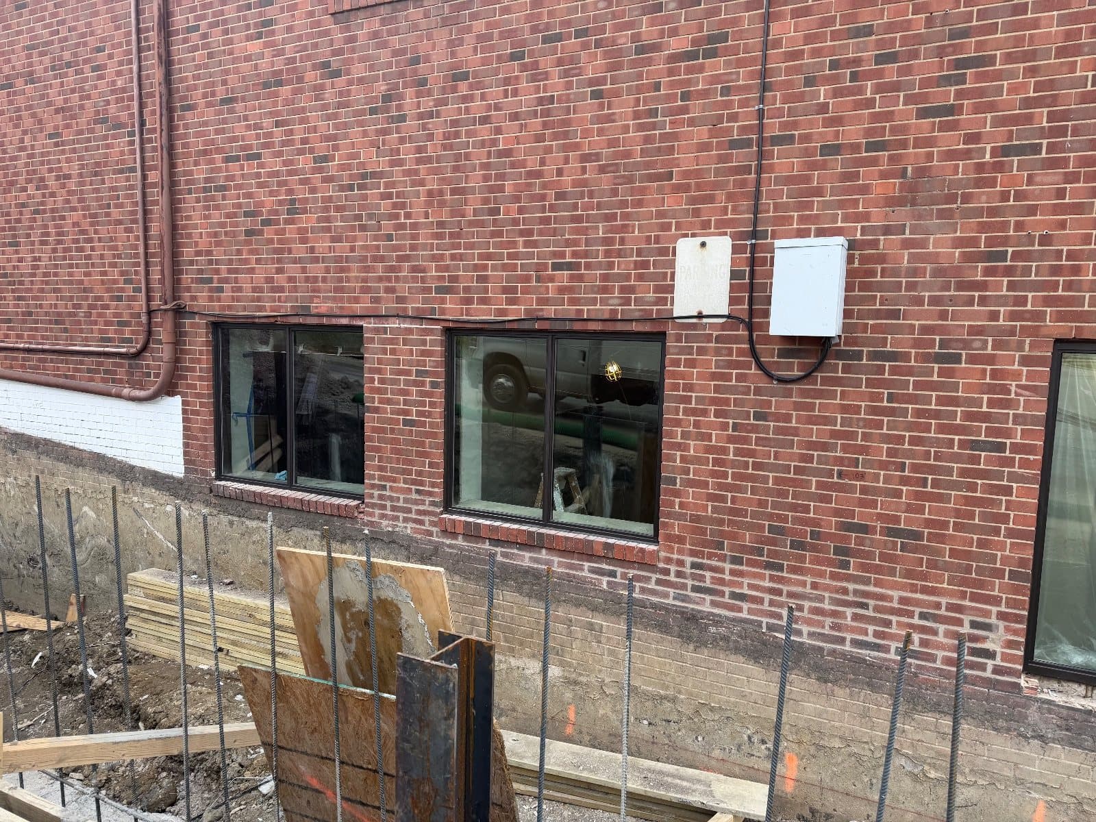 Two ground-floor picture windows in dark aluminum frames set in the brick wall, the excavated foundation and rebar in front of them.