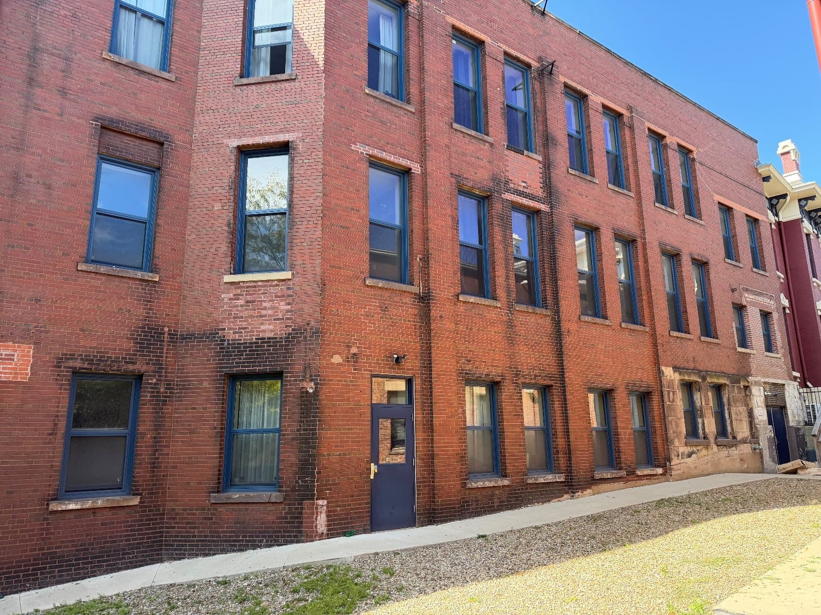 The long red brick elevation seen at an angle, tall windows in dark blue frames running its length, a red and cream building at the far corner.