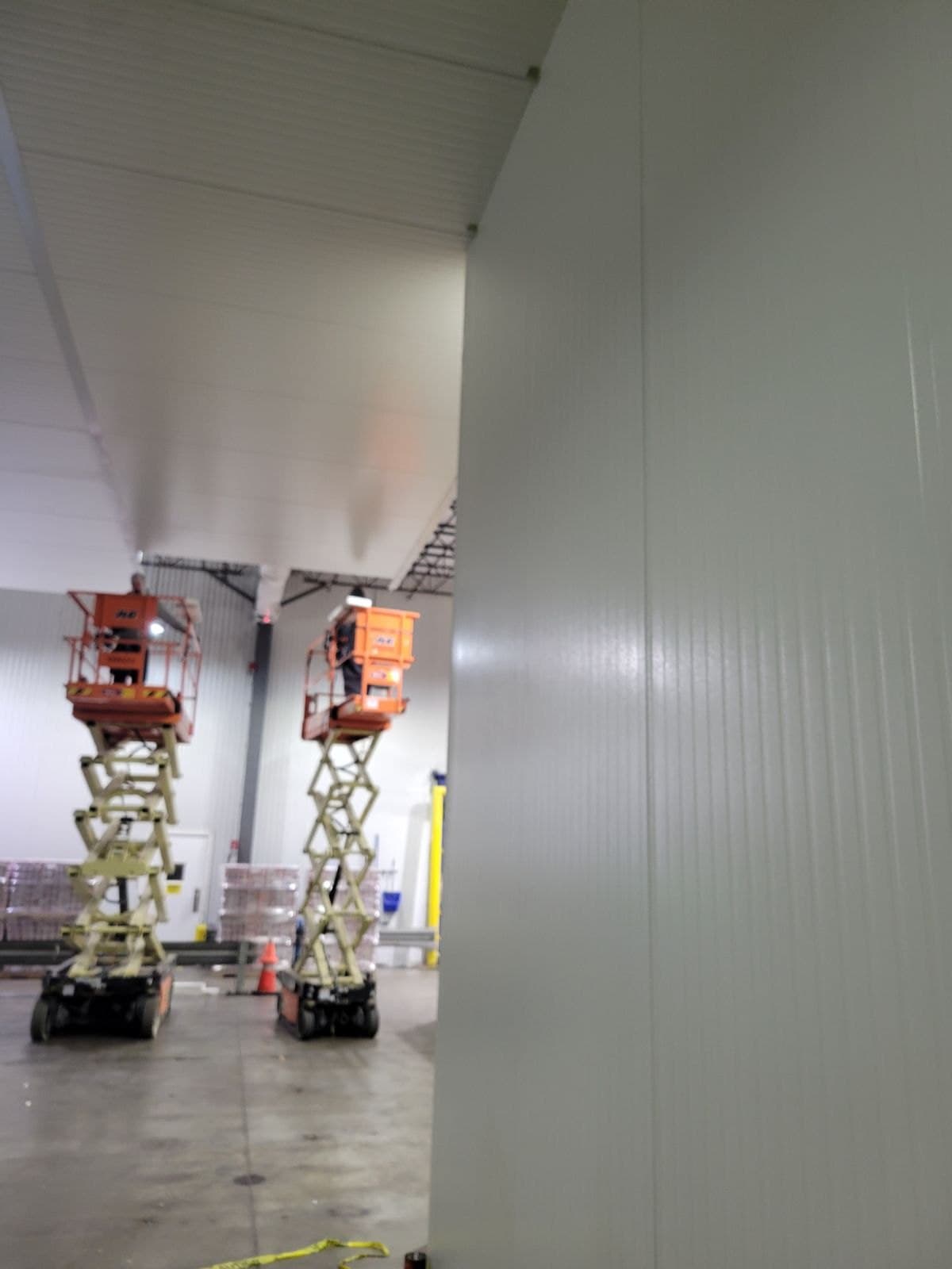 A white insulated panel wall in the foreground, two scissor lifts beyond it with workers at the ceiling.
