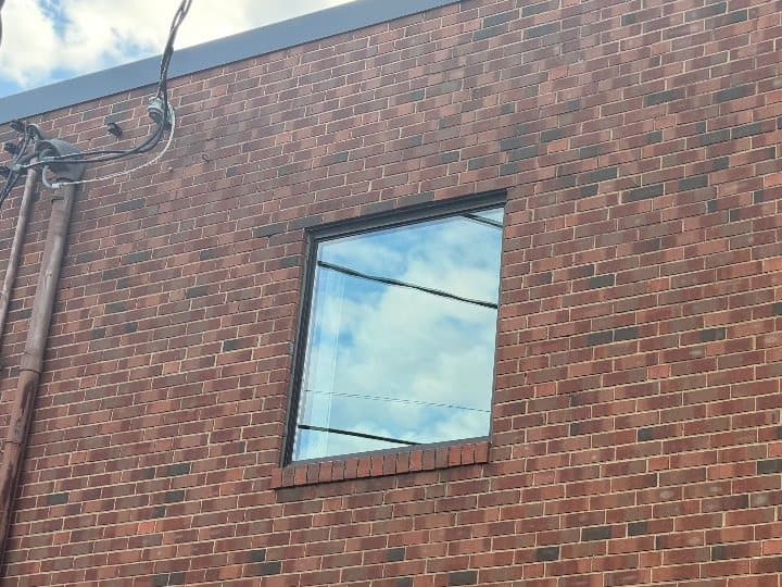 A single square picture window in a dark aluminum frame high on a red brick wall, sky reflected in the glass.