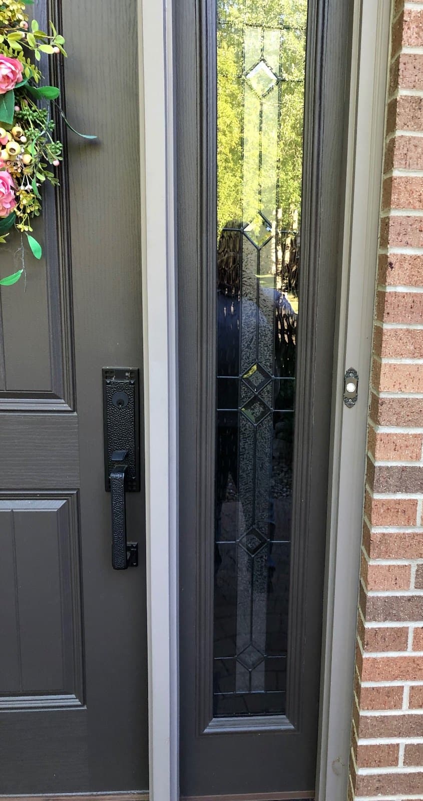 Close view of a gray entry door edge with a handleset, beside a tall decorative leaded sidelite set in brick.