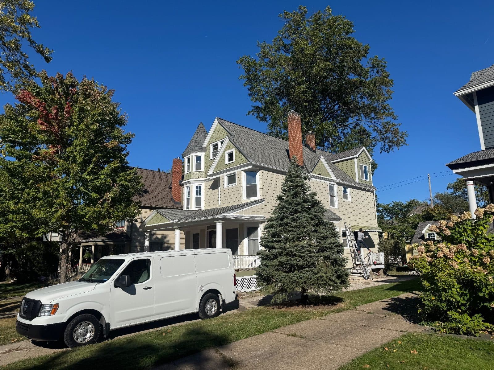 The Victorian seen from down the street, sage and cream siding and white window frames on every floor, a ladder against the side porch and a white work van at the curb.