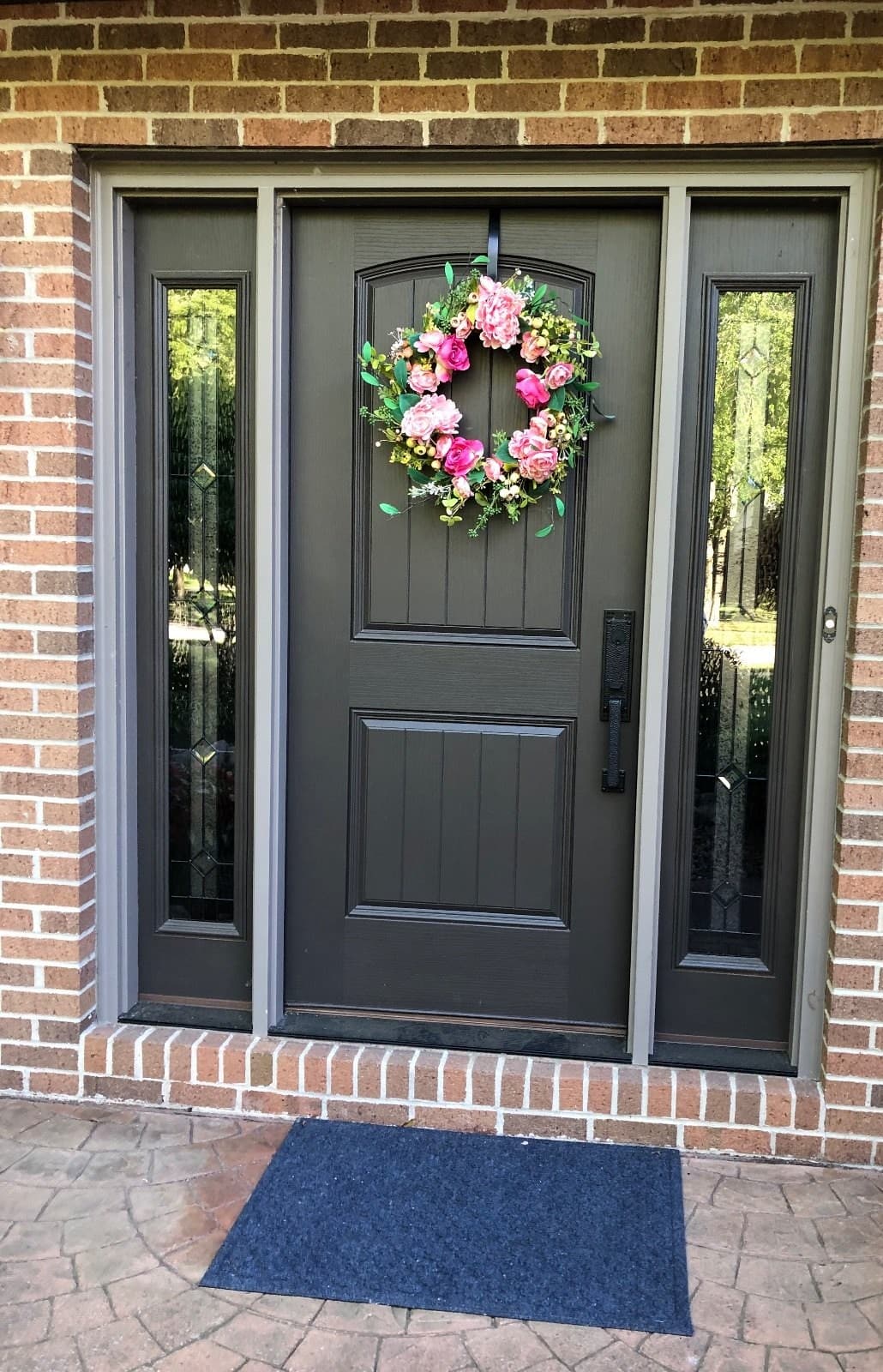 A gray entry door in place on a brick house, a flower wreath on the door and a narrow leaded sidelite each side.