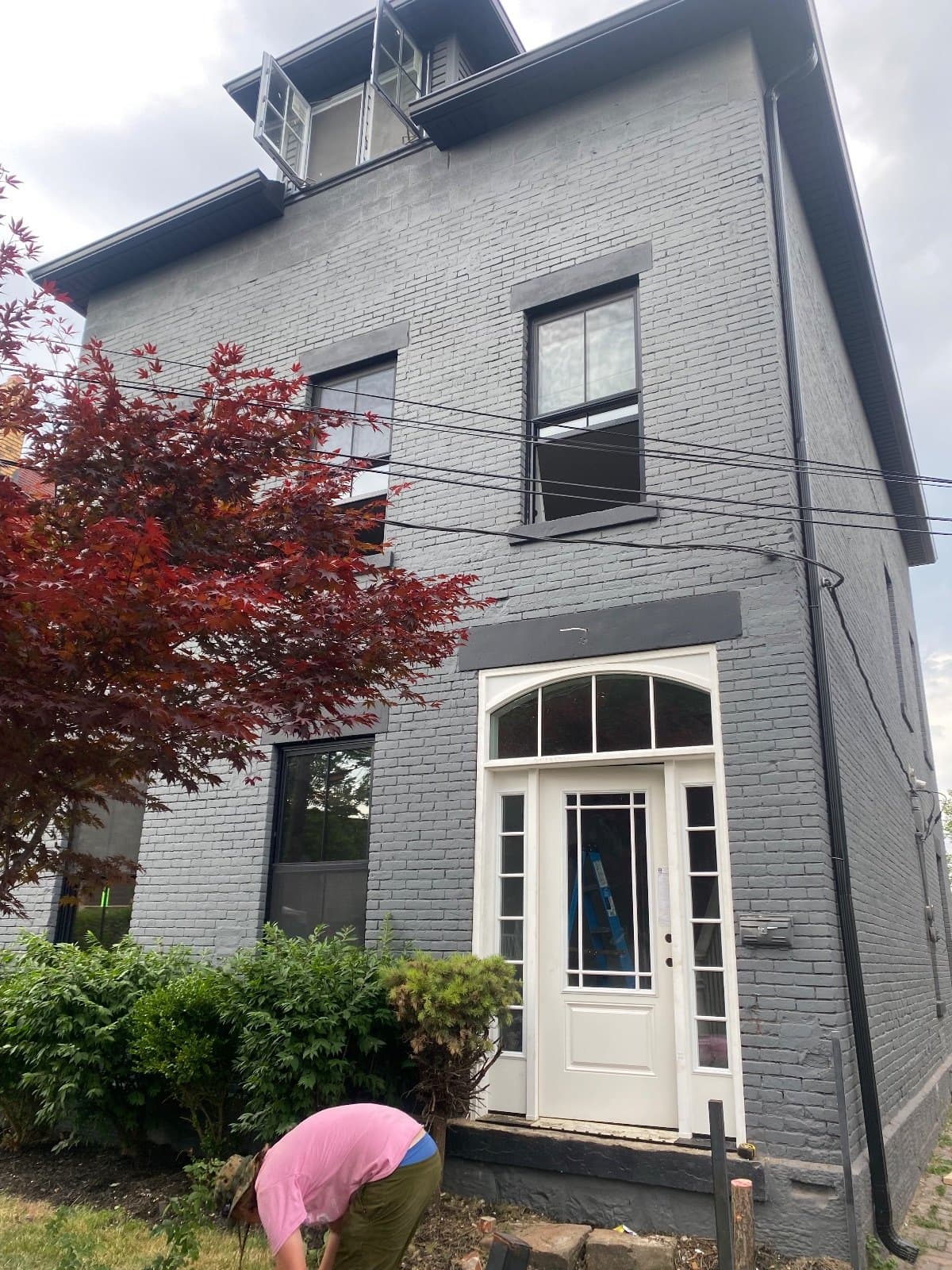 A white entry door with an arched transom above it, in place on a gray brick house behind a red maple.