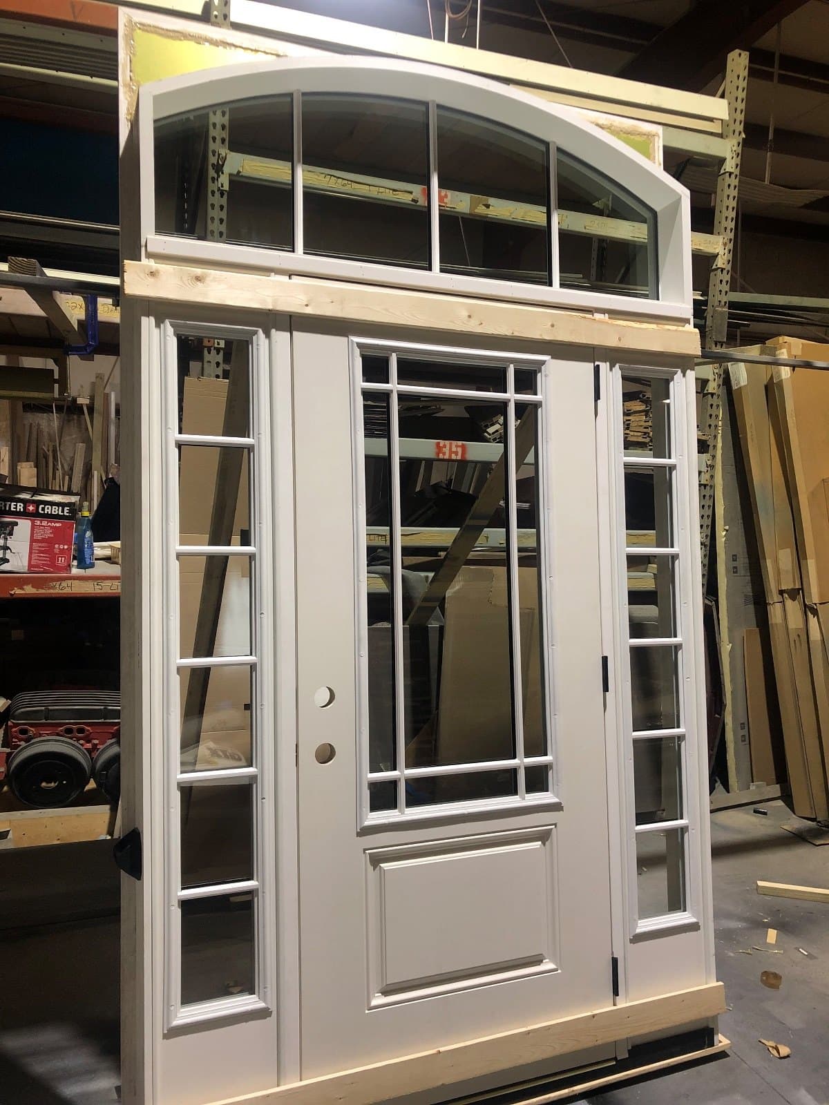 A white entry door with two sidelites and a wide arched transom, braced upright in the shop.