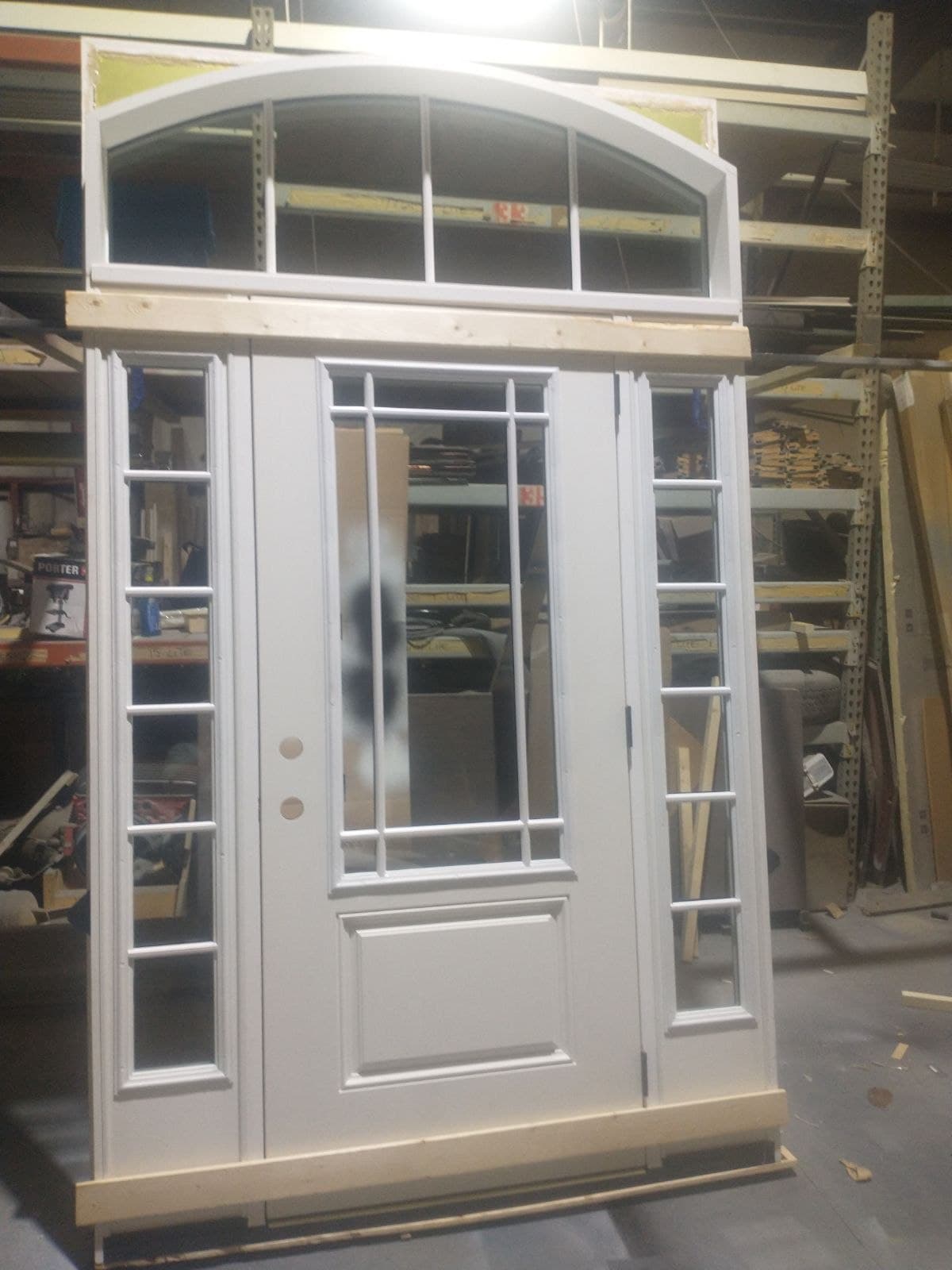 A custom white entry unit standing in the shop, grid sidelites either side and a segmental arched transom above, still on its shipping blocking.