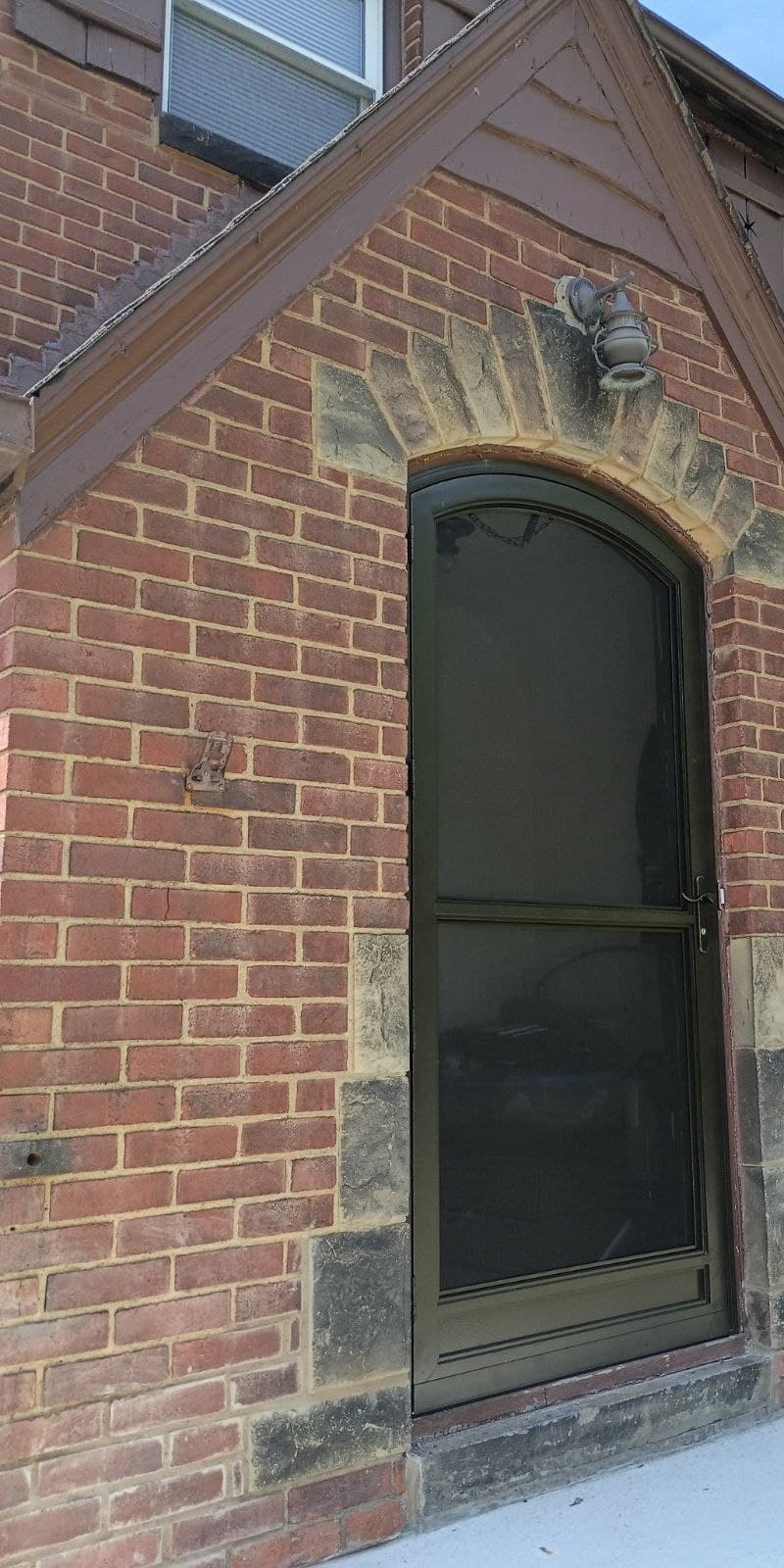 A radius-topped storm door installed in an arched brick opening, cut stone voussoirs over it and a lantern on the brick.