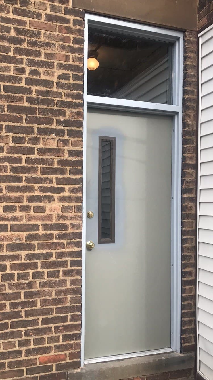 A gray commercial door with a narrow vertical vision lite, set in a metal frame with a glass transom above, in a brick wall.