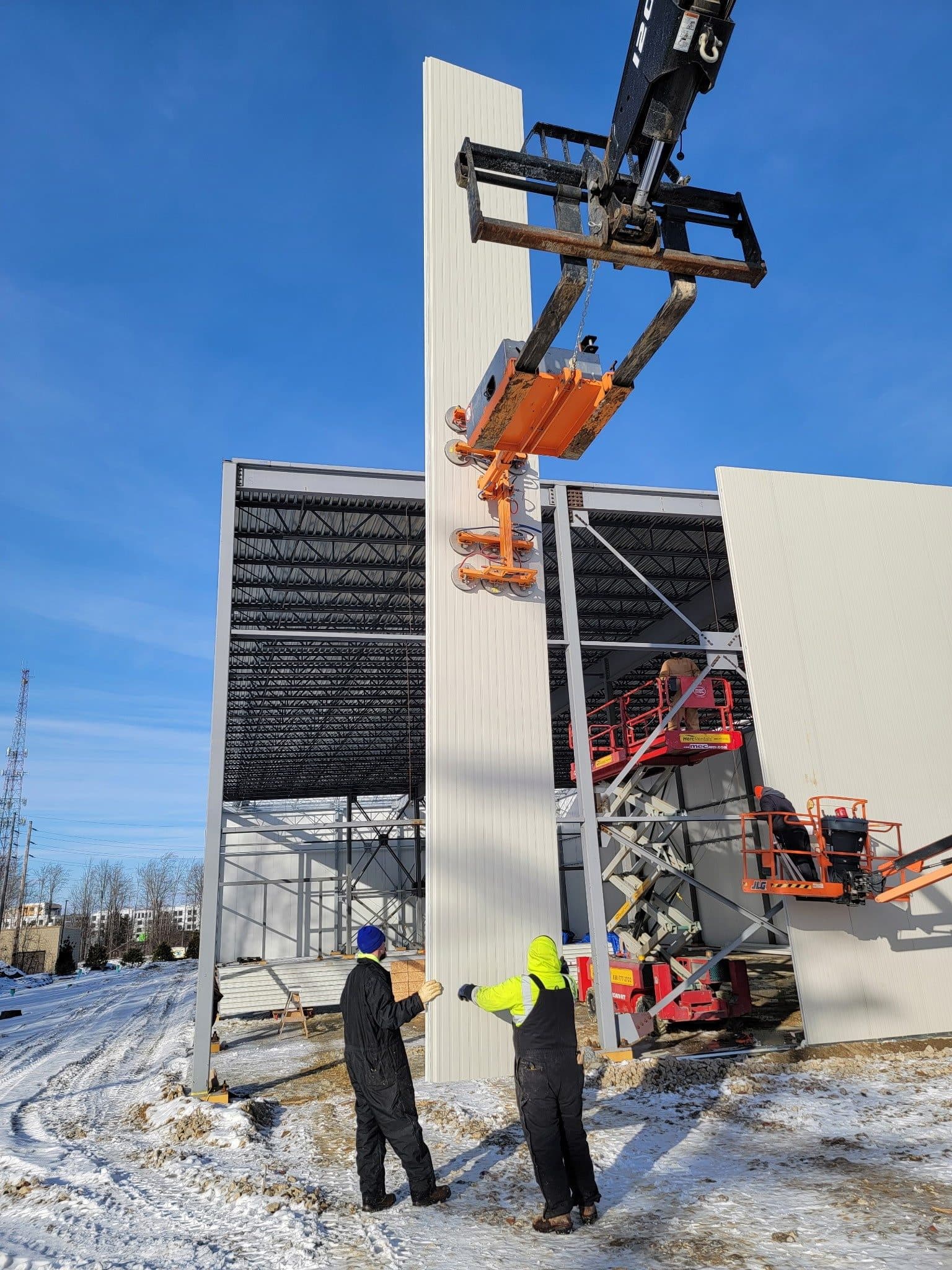 Two 32-foot insulated metal panels being lifted into place against a blue winter sky, installers below.