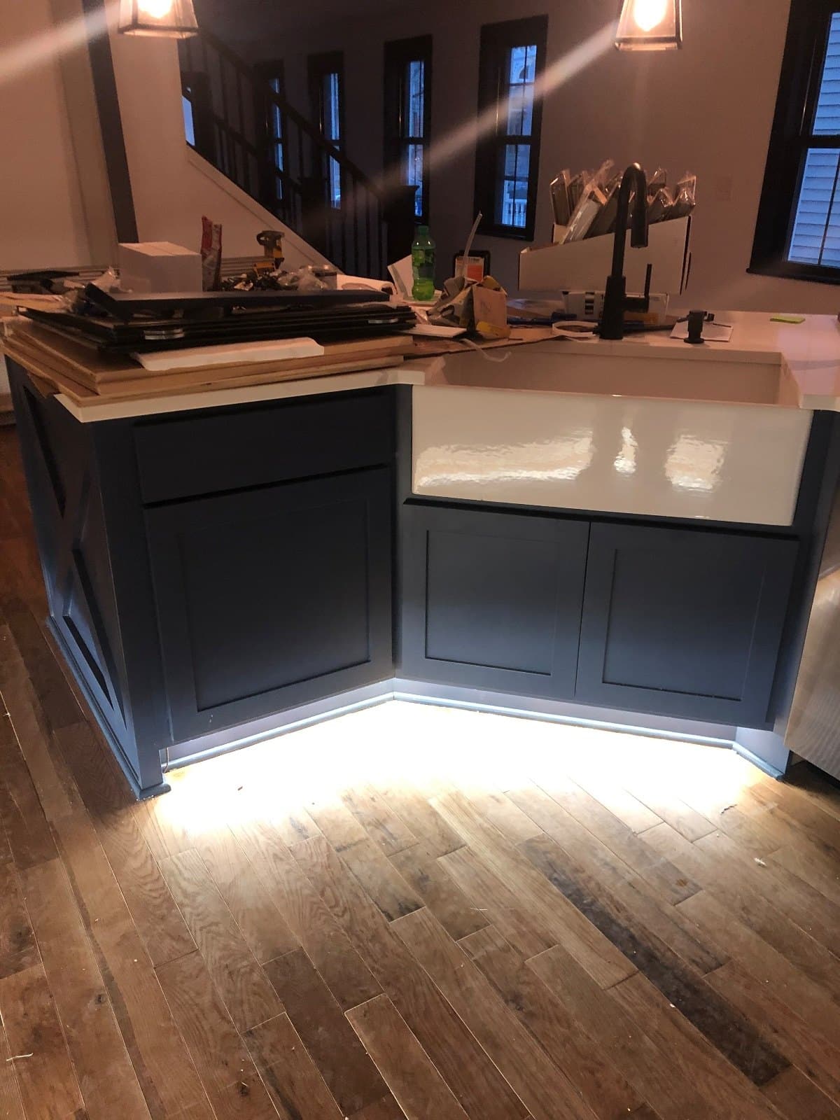 A farmhouse sink and dark island corner with materials stacked on the counter, hardwood down around them.