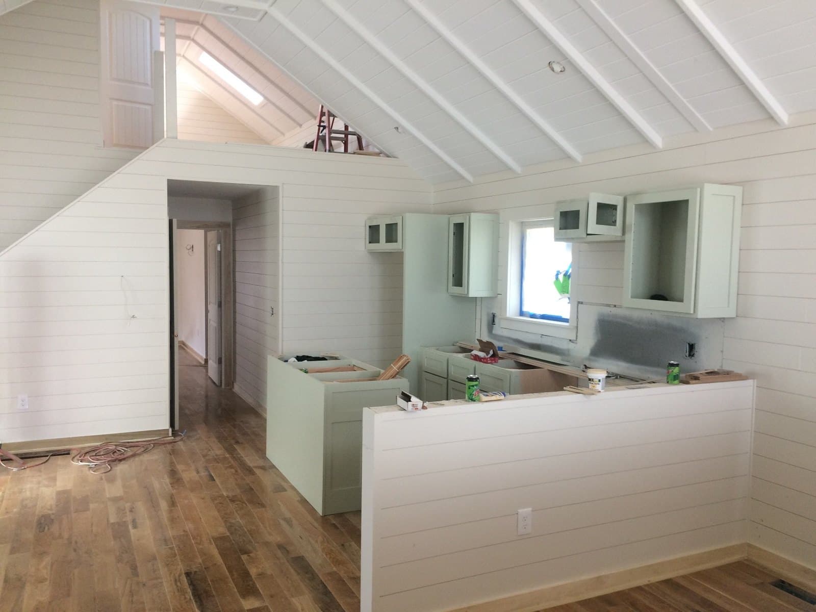 An unfinished kitchen under a vaulted white ceiling, doors off the uppers and a cord across the hardwood.