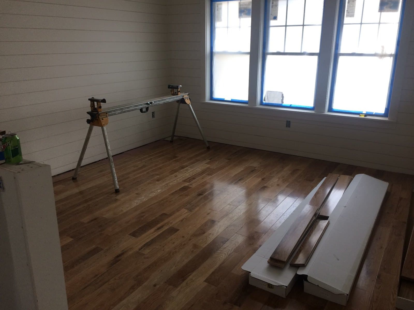 A miter saw on a stand and lengths of trim stock lying on a finished hardwood floor by three windows.