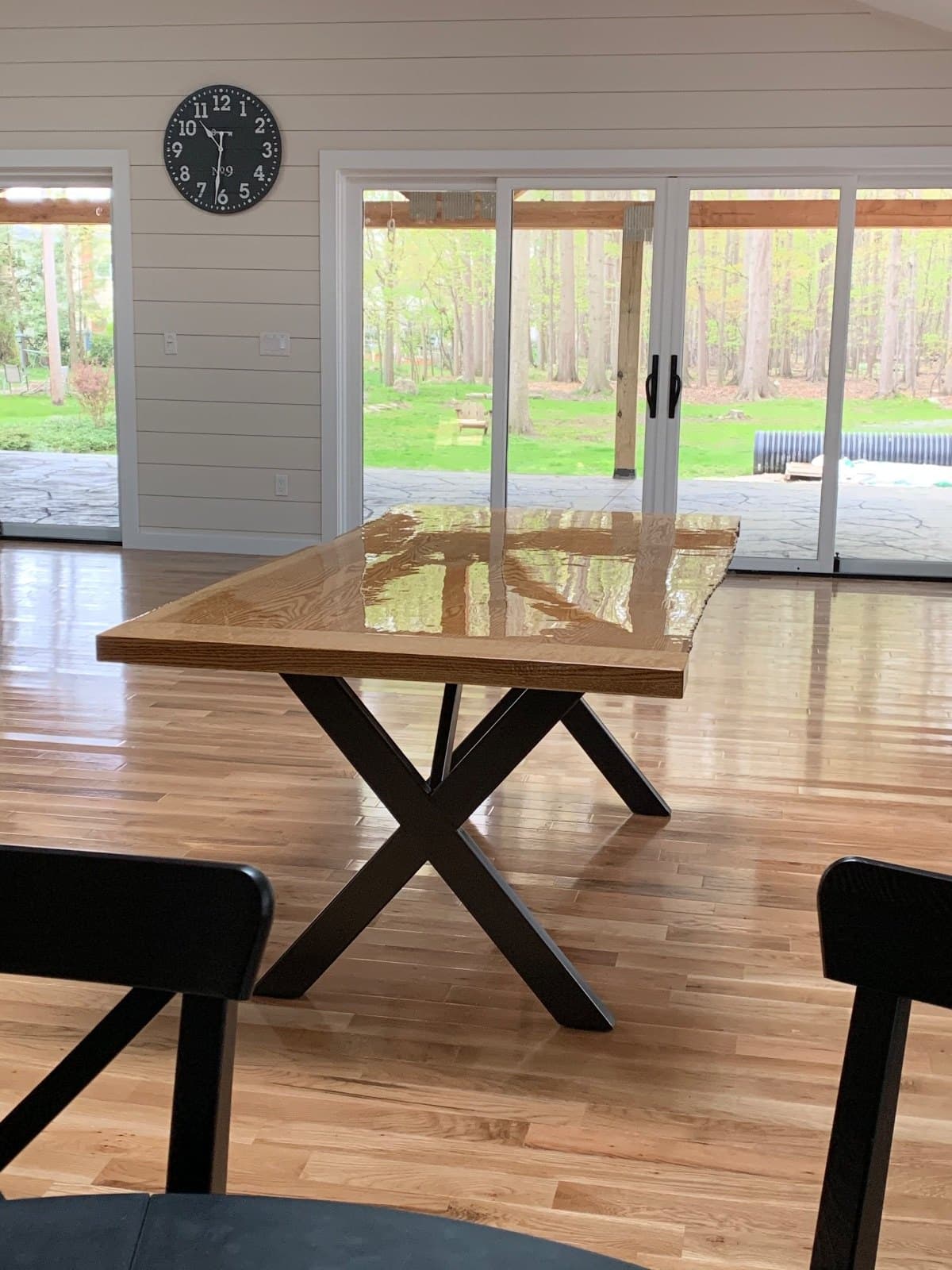 A live-edge dining table on a hardwood floor, a wall clock behind it and sliding glass doors onto woods.