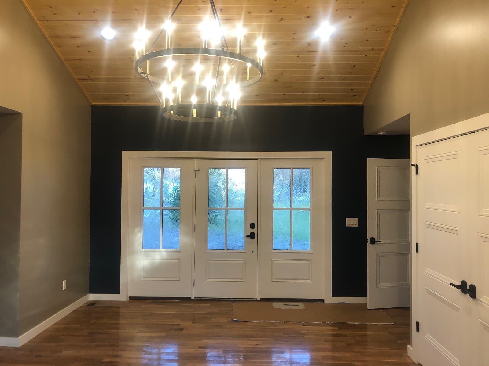 An entry with double glass doors under a ring chandelier, dark walls and a plank ceiling above hardwood.