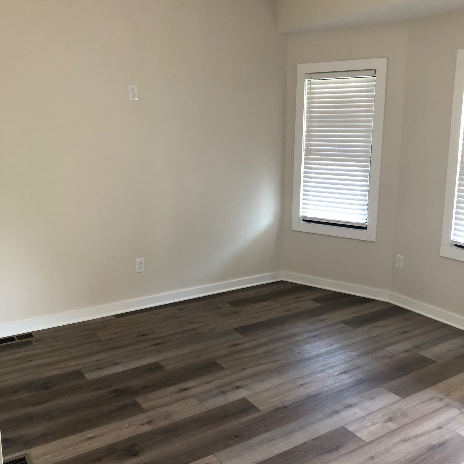 An empty room floored wall to wall in gray-brown vinyl plank, a blinded window on the far wall.