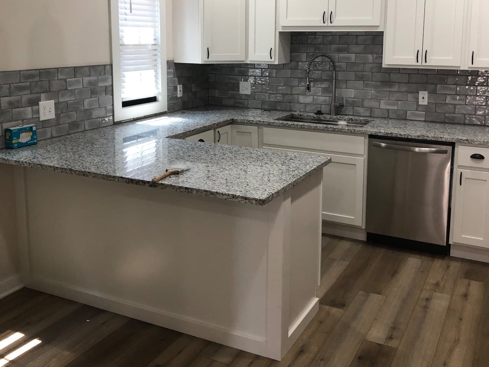 Vinyl plank running across a kitchen floor past a granite peninsula, gray brick backsplash behind.