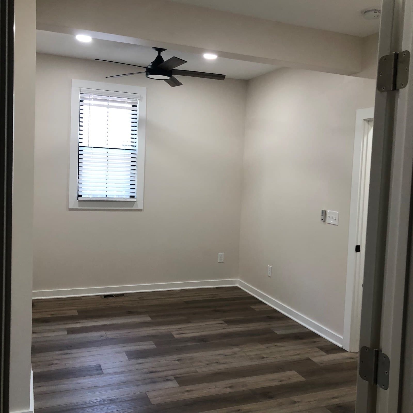 A living room under a ceiling fan with a blinded window and a doorway, vinyl plank throughout.
