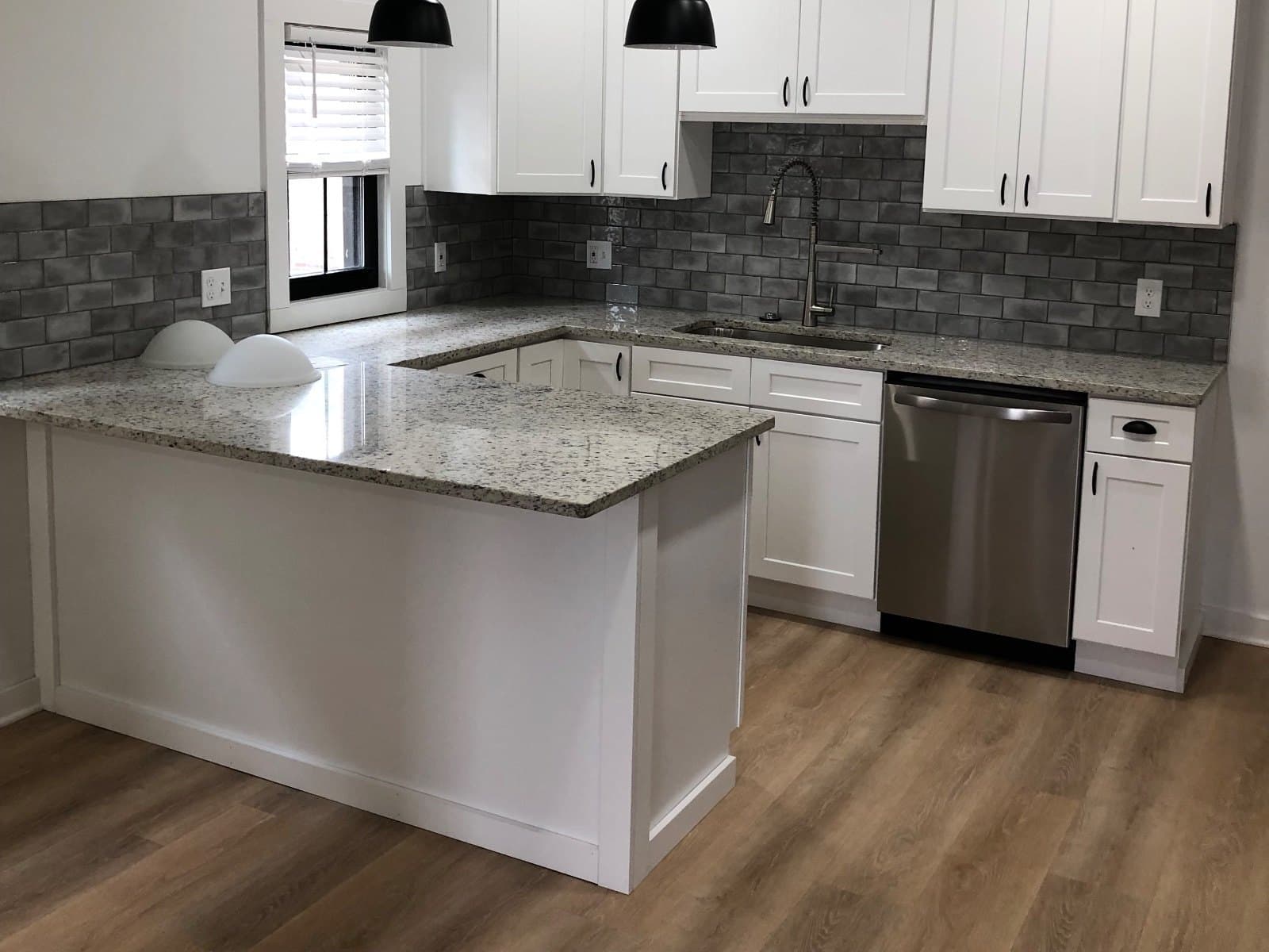 A kitchen with granite counters, gray brick backsplash and black pendants over vinyl plank.