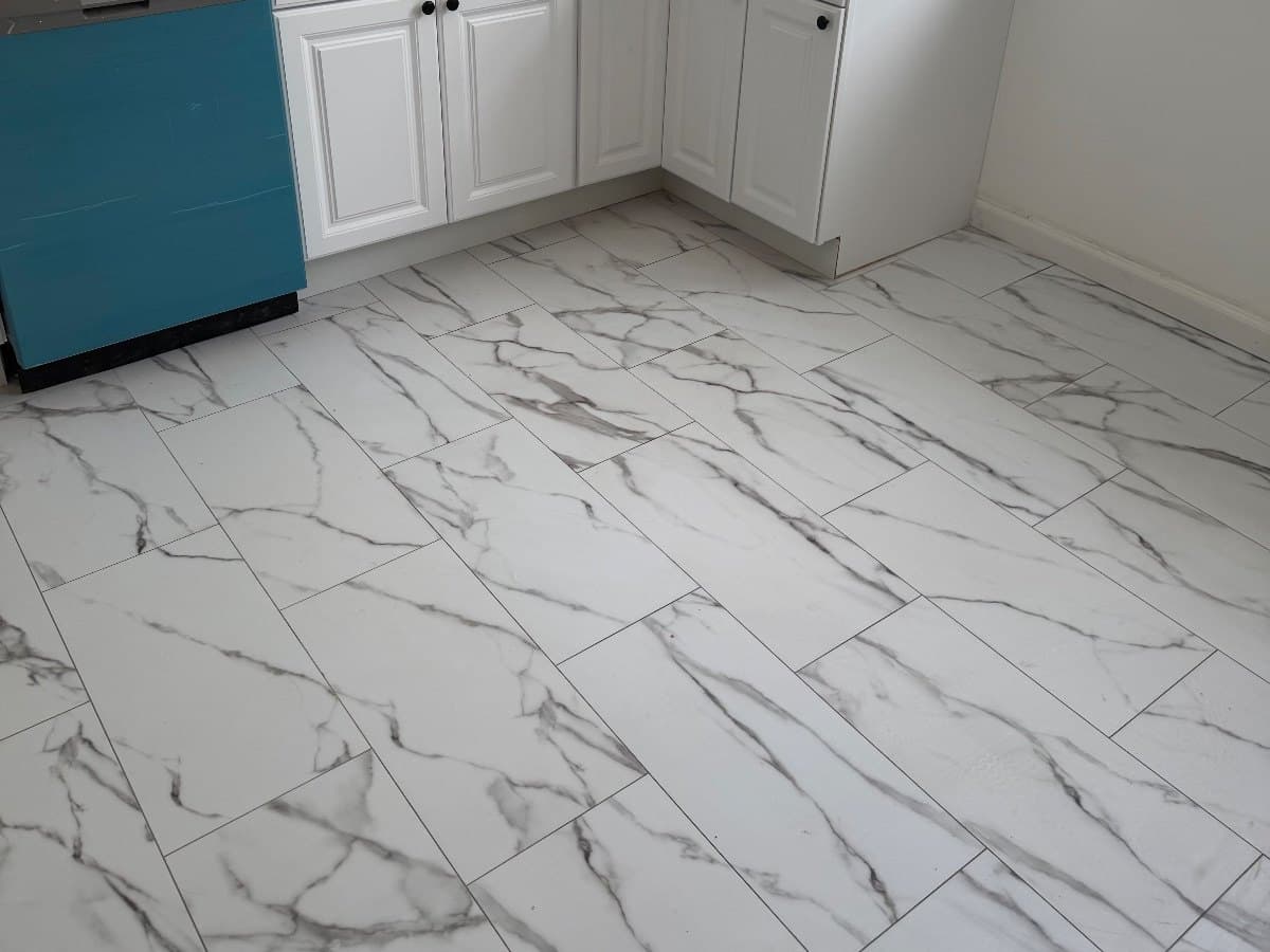 A kitchen floor in wide vinyl plank printed to look like veined marble tile, running past a teal dishwasher panel toward a baseboard corner.