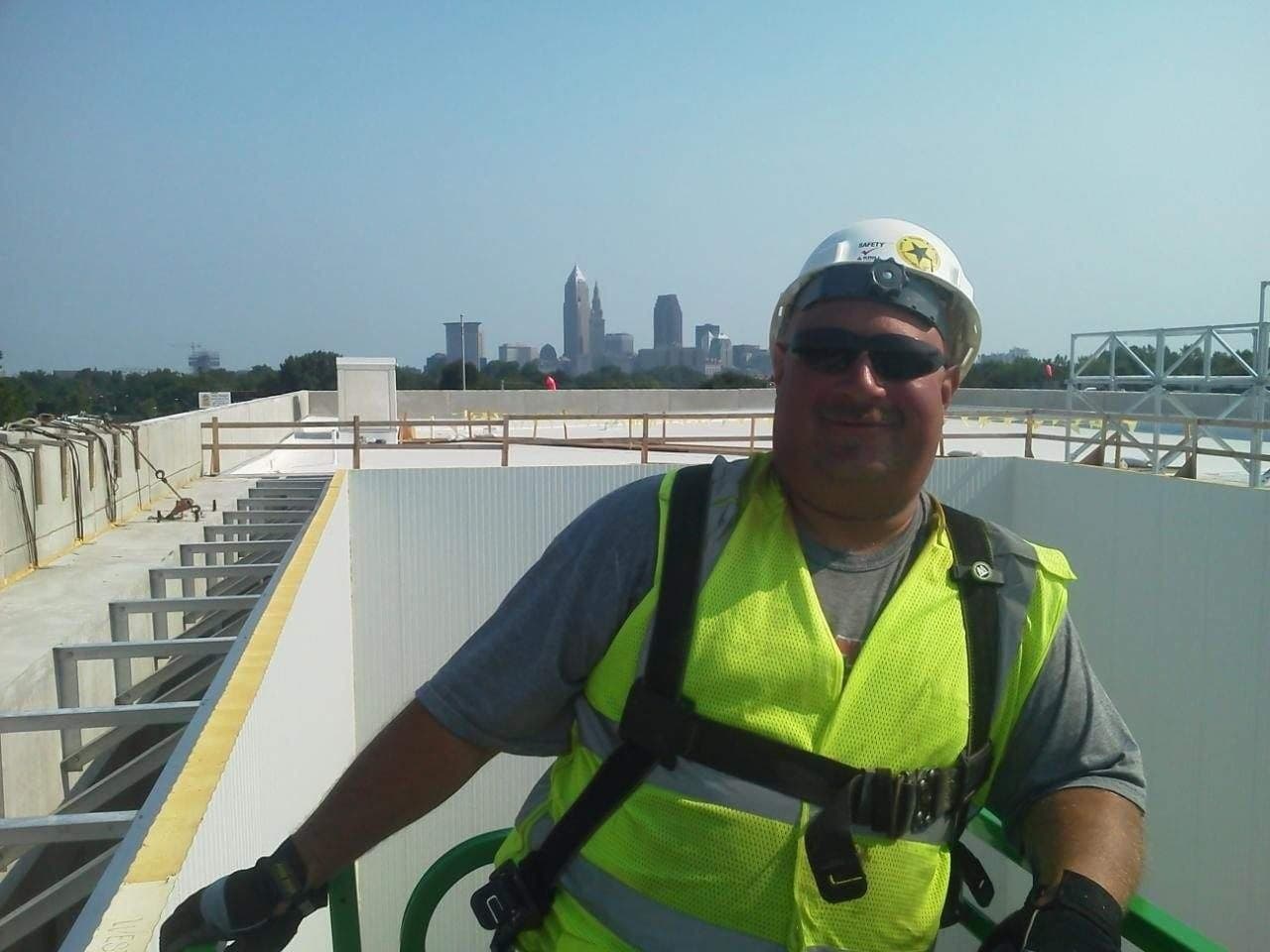 An installer in a hard hat, safety harness and high-visibility vest on a lift beside a white insulated panel wall on a roof, the Cleveland skyline behind.