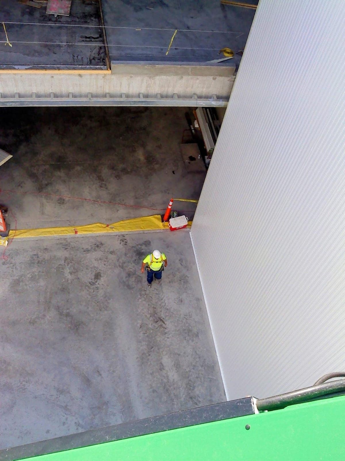 Looking down from a lift along a tall white insulated panel wall to an installer in a hard hat and high-visibility vest on the floor below.