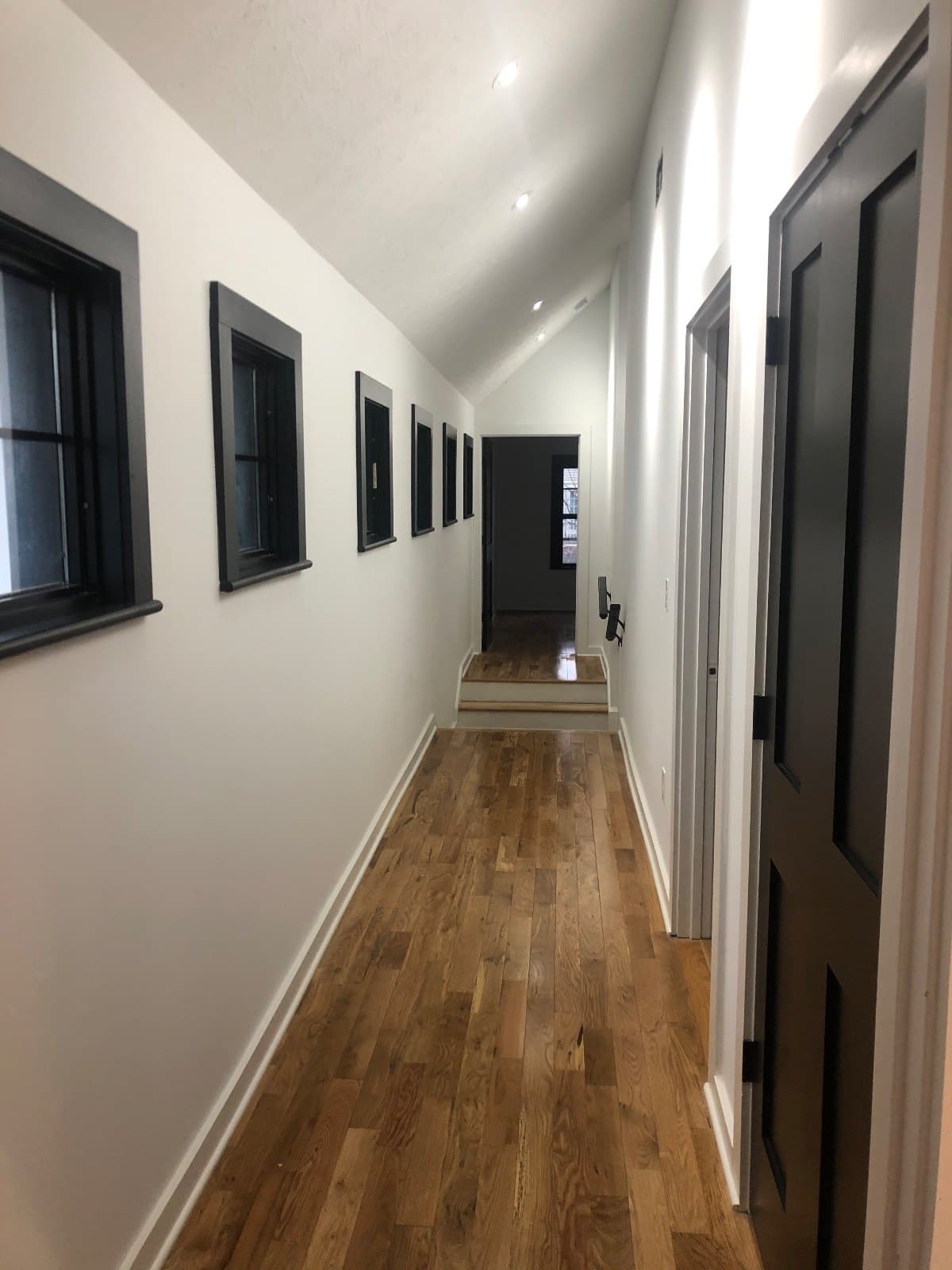 A hallway in white walls and wood floor, tall dark-framed windows down one side.