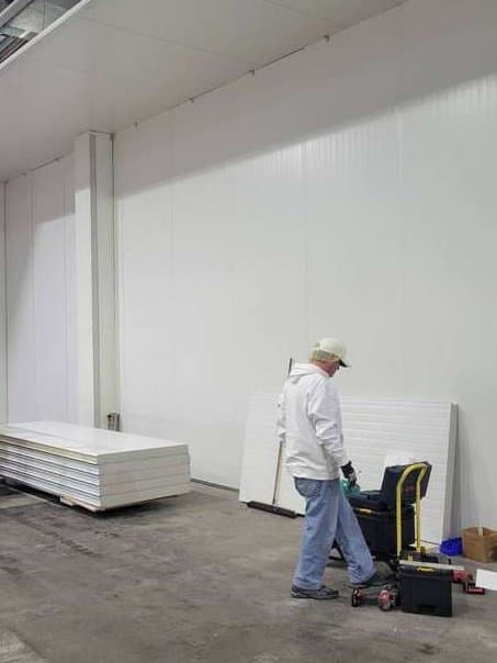 A long white insulated panel wall inside a plant, panels stacked on the floor and an installer at his tool cart.