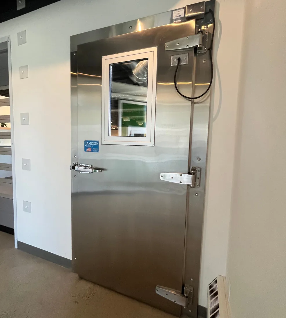Stainless steel Jamofoam II swing door with a vision window, seen at an angle with its hinges